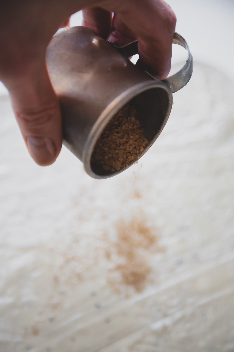 hand sprinkling cane sugar from an old aluminium measuring cup on a sheet of cinnamon bun dough