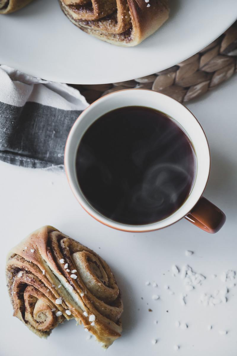 steaming hot black coffee and a delicious vegan cinnamon bun on a white table next to a plate of buns