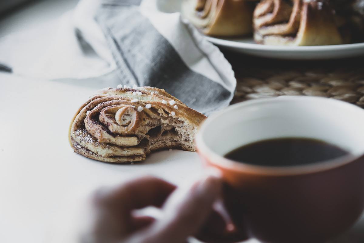 hand holding a cup of coffee while focus is on a cinnamon bun that's half eaten