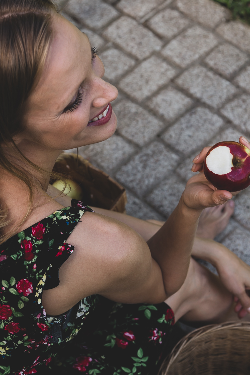 A smiling woman wearing a dress is sitting outdoors and has just taken a bite of a fresh apple.