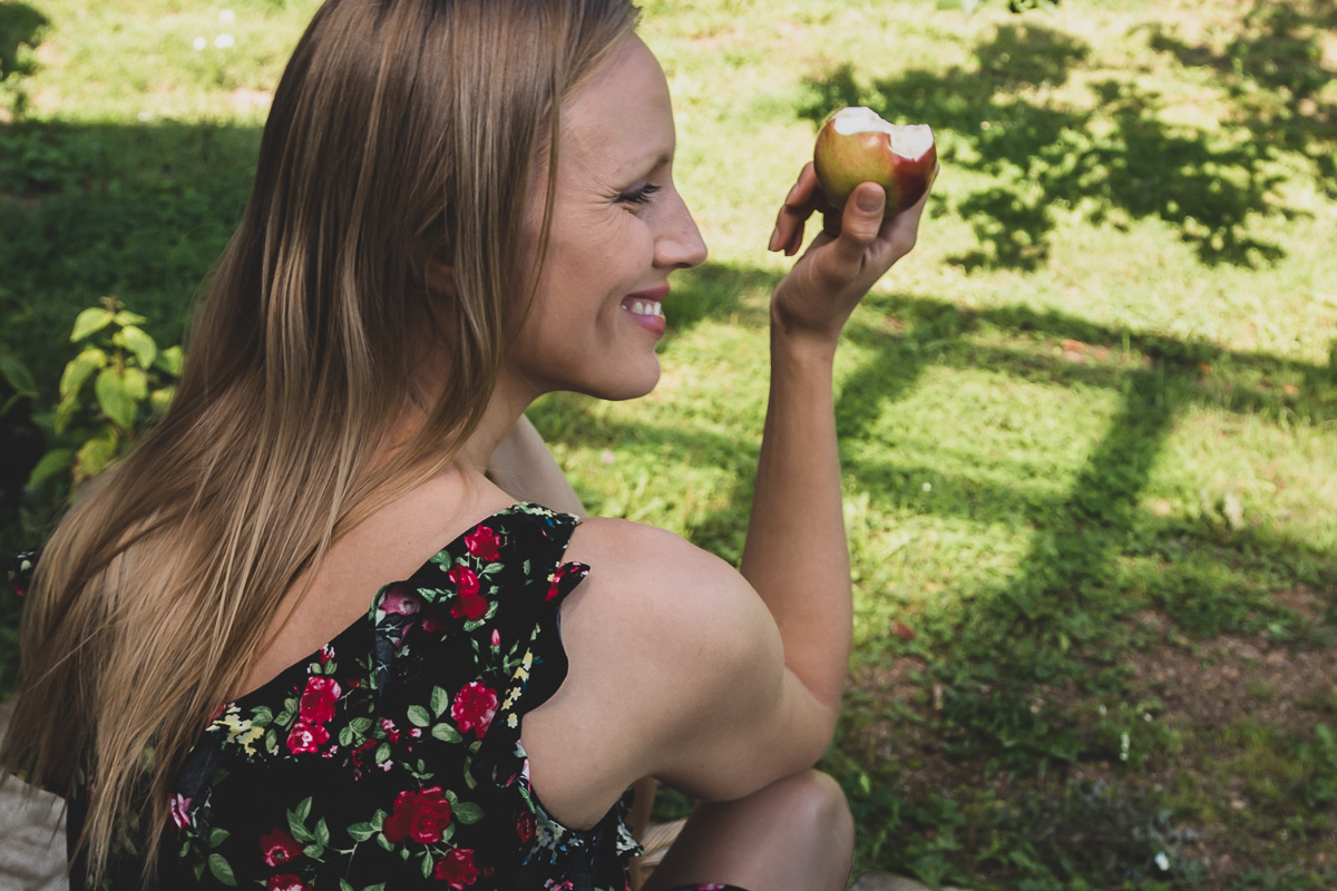 A smiling woman wearing a dress is sitting outdoors and has just taken a bite of a fresh apple