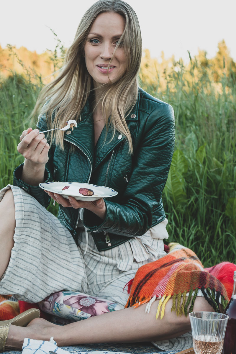Woman in green faux-leather jacket is eating dessert while having a sunset picnic on a green field