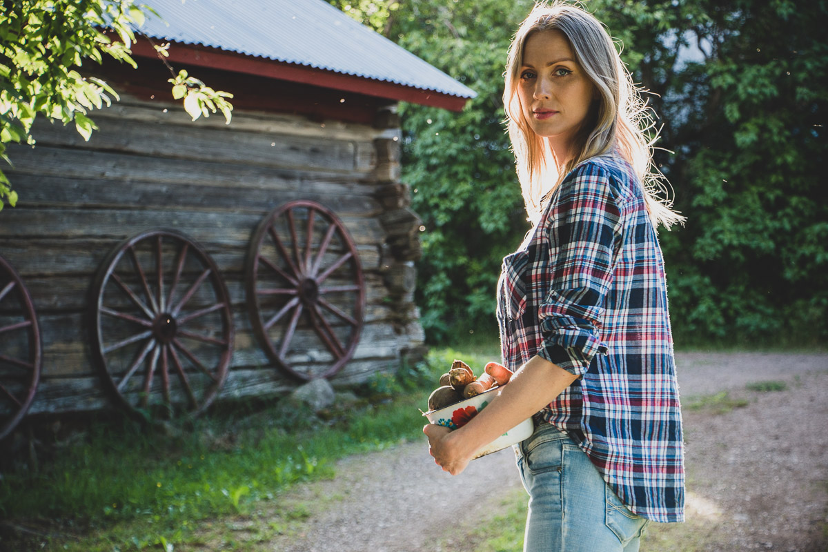 A woman holding a bowl of root vegetables outside a country style shed.