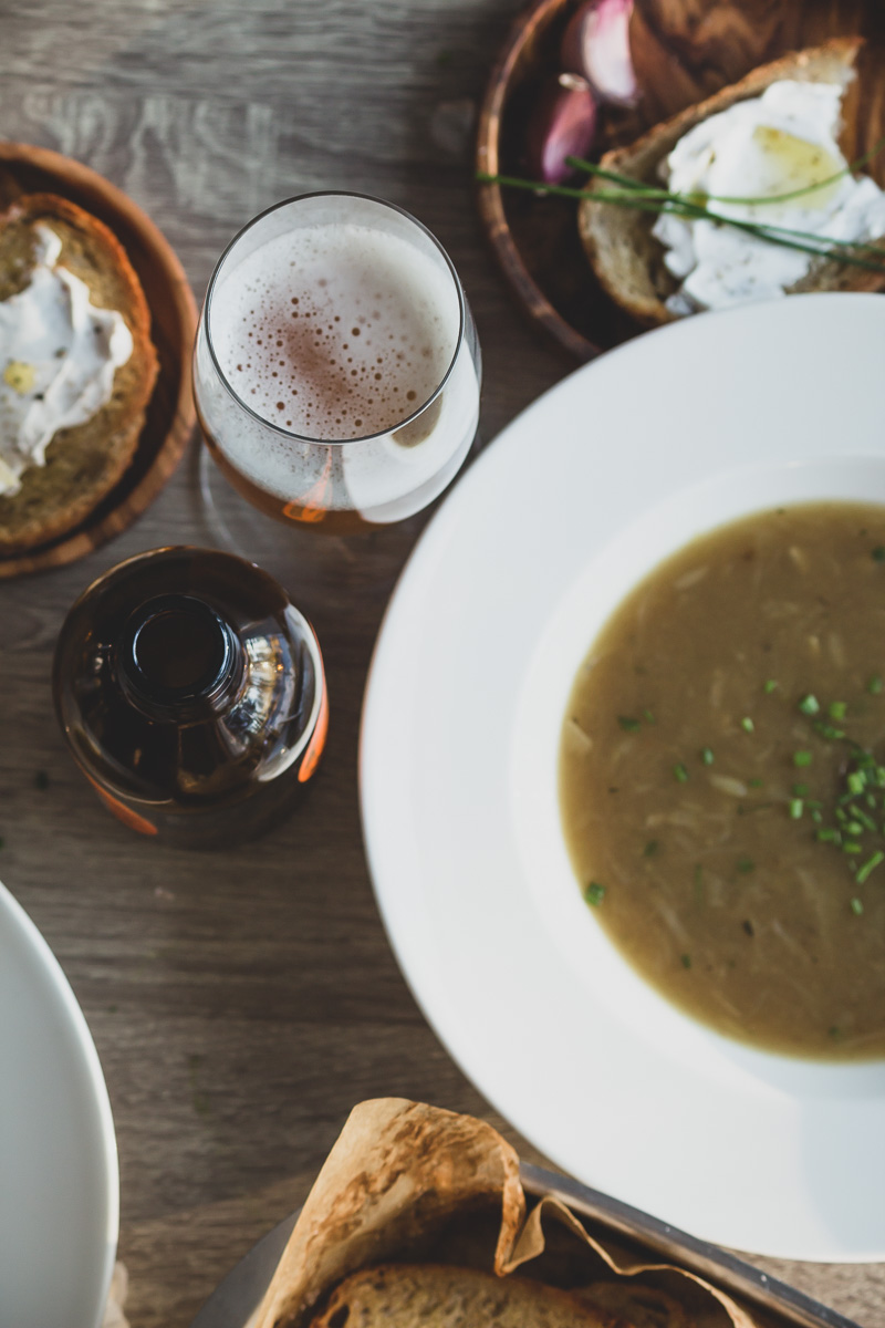 A plate of onion soup, crunchy slice of bread with some vegan cream cheese and chives on top of it, and a glass of beer.
