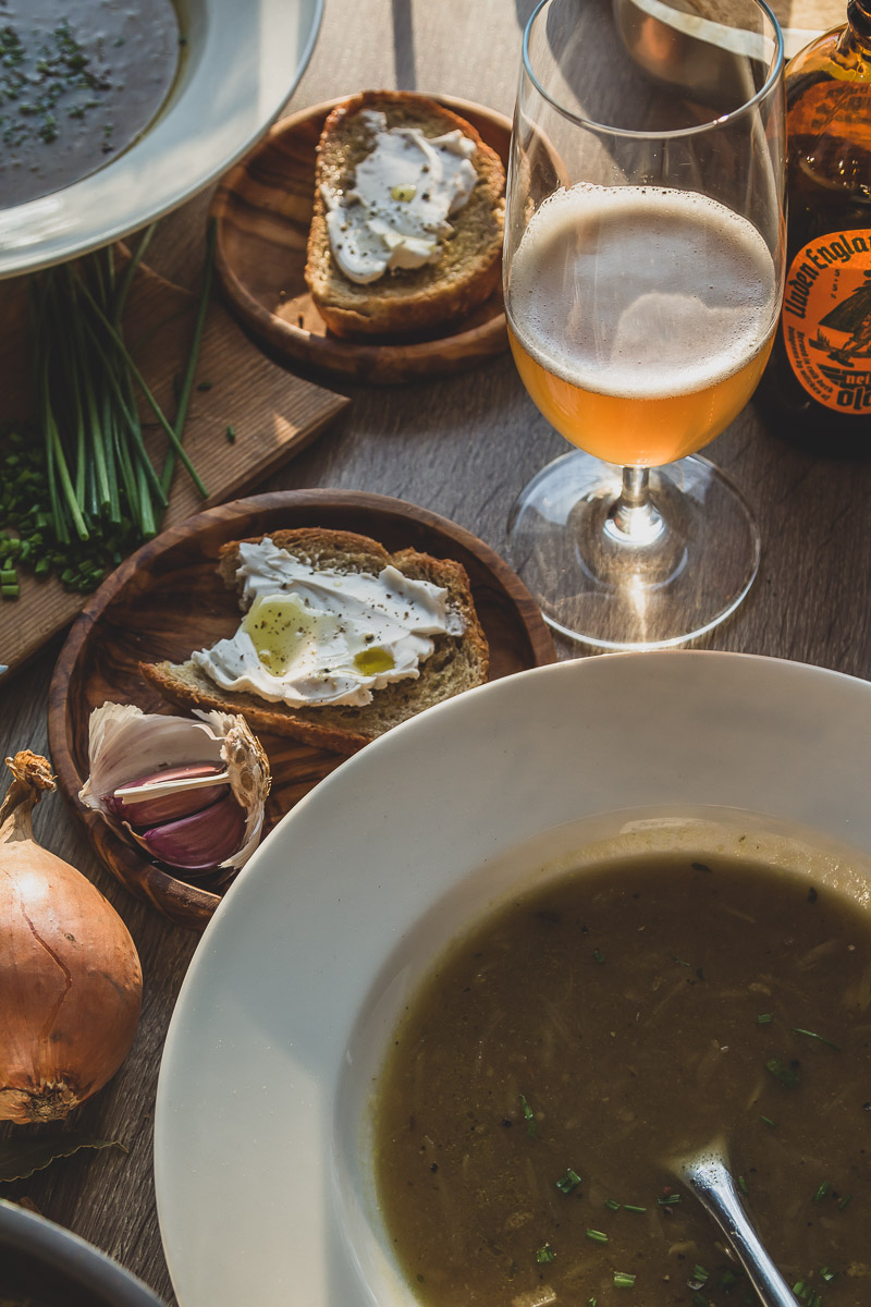 A plate of onion soup, crunchy slice of bread with some vegan cream cheese and chives on top of it, a beer glass and onions, spices scattered around the plate on the table.