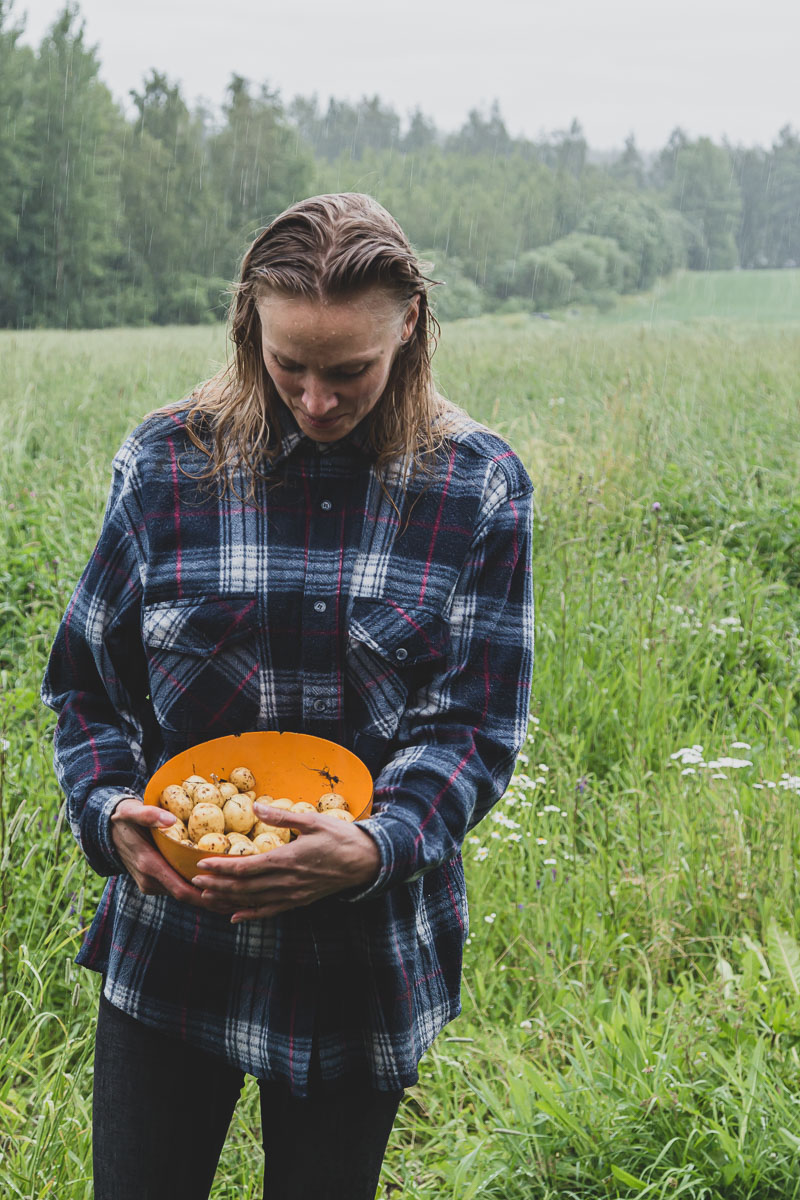A woman wearing a blue flannel shirt is standing in the rain, on a field looking down to a bowl of freshly harvested potatoes she is holding in a n orange metal bowl.