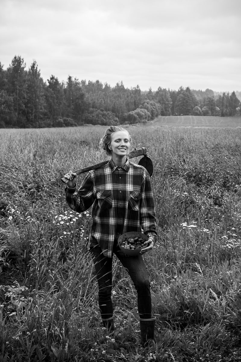 a black and white portrait of a woman, wearing a flannel shirt, holding freshly harvested potatoes on a bowl and a pickaxe. She looks proud and is smiling in the rain.