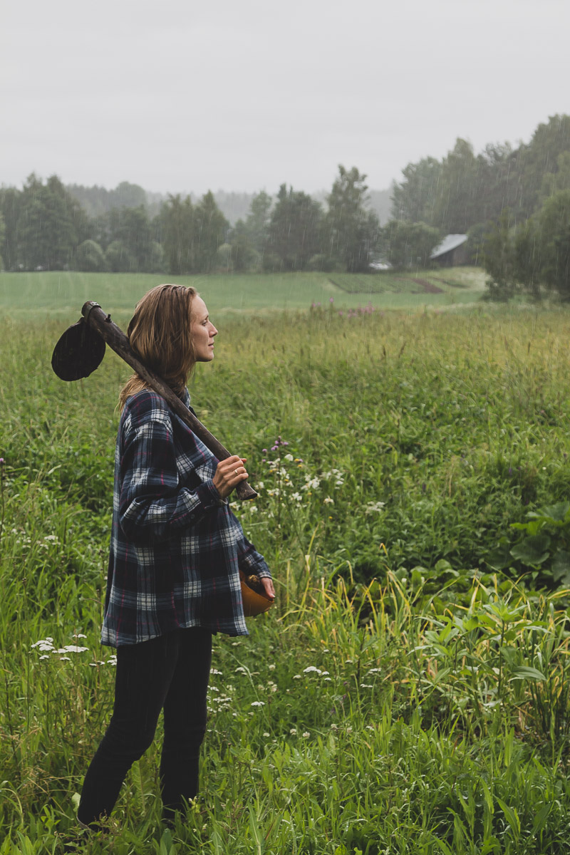 A woman stands in the rain on a side of a field holding a bowl of freshly harvested potatoes.